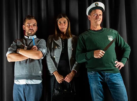 Ein Polizist, ein Kommissar und eine Kommissarin stehen nebeneinander vor einem Vorhang