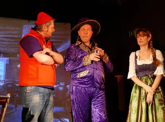 Ein Mann mit roter Mütze und Weste, einer im lila Samtanzug und eine Frau im Dirndl